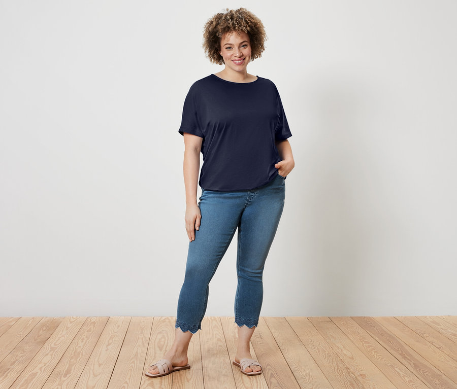 Une femme aux cheveux bouclés porte un t-shirt bleu marine, un jean bleu avec un ourlet ondulé et des sandales.