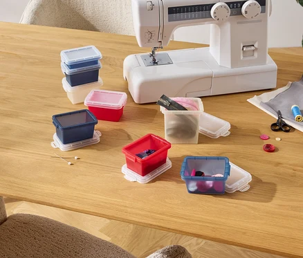 Sur une table en bois se trouvent une machine à coudre et plusieurs boîtes de rangement colorées contenant des accessoires de couture tels que des boutons, du fil et des fermetures éclair.