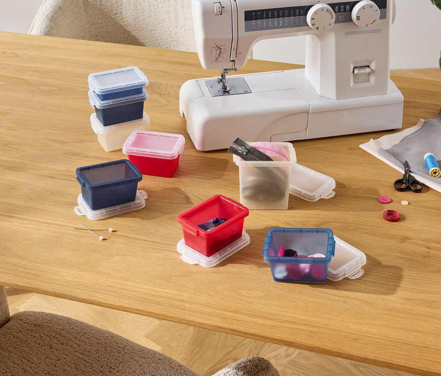 Sur une table en bois se trouvent une machine à coudre et plusieurs boîtes de rangement colorées contenant des accessoires de couture tels que des boutons, du fil et des fermetures éclair.