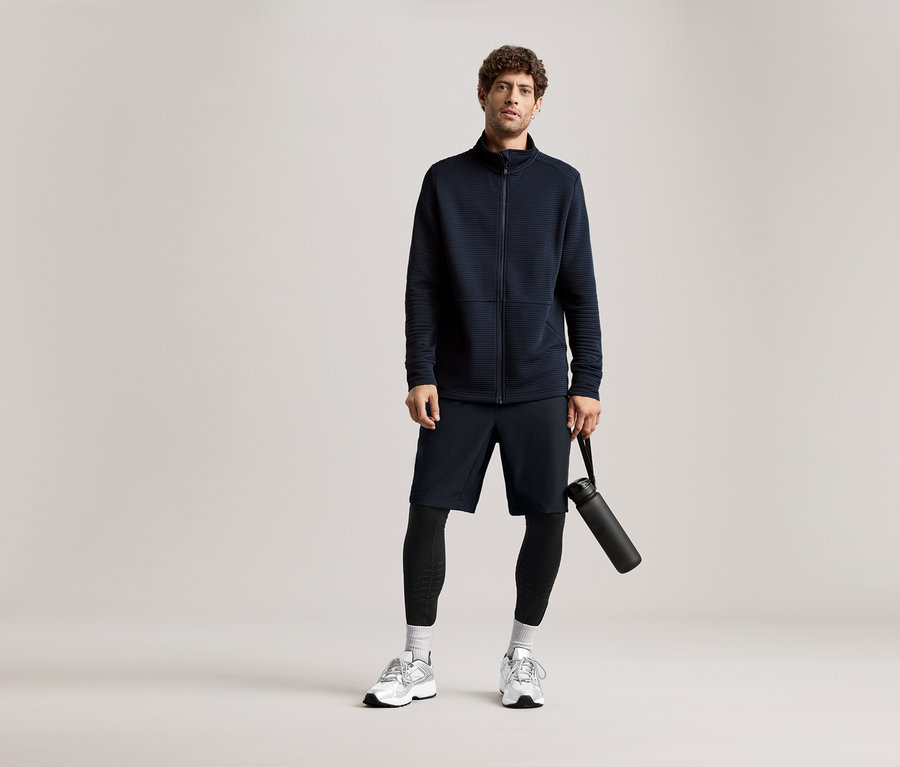 Homme en veste bleu marine, short et leggings, tenant une bouteille d'eau noire.