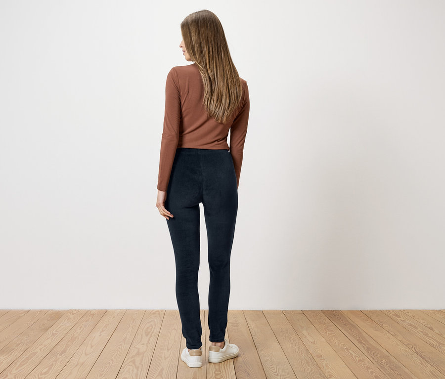Femme portant un haut marron et un legging bleu marine, vue de dos, devant un mur blanc et un plancher en bois.