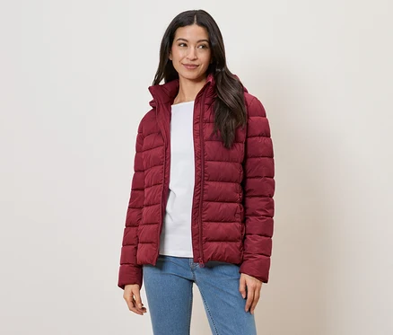 Une femme aux longs cheveux foncés porte une doudoune rouge, un t-shirt blanc et un jean sur un fond clair.