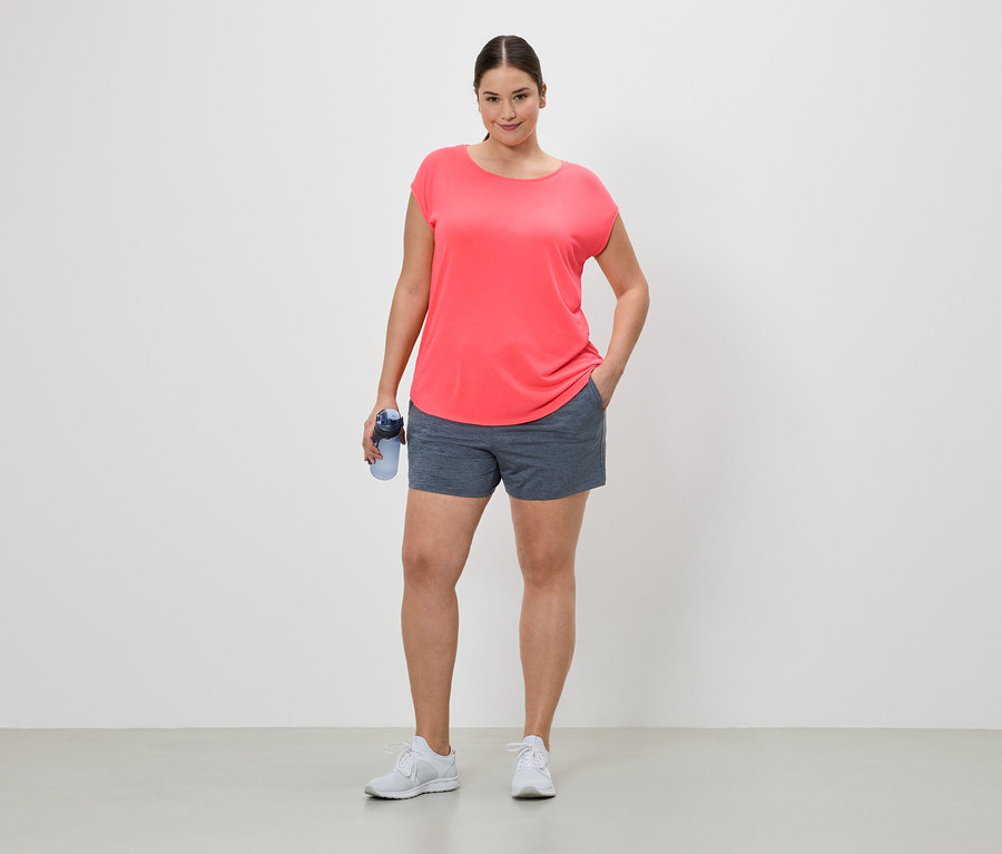 Femme en tenue de sport avec une bouteille d'eau, debout sur un fond blanc. Elle porte un t-shirt corail, un short et des baskets blanches.