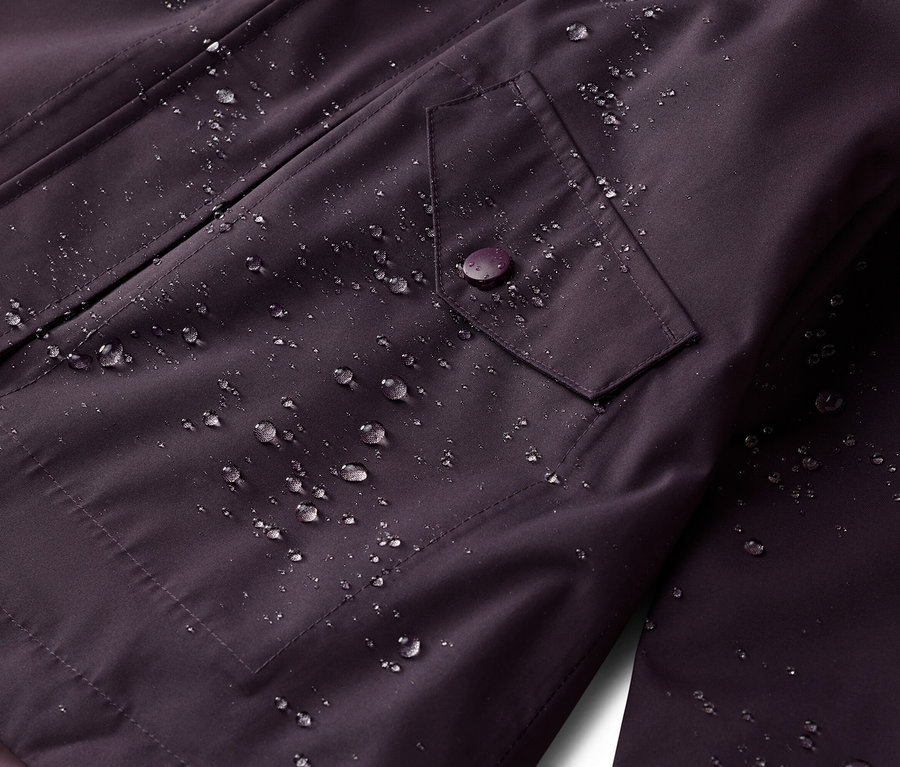 Gros plan sur une veste aubergine imperméable avec une poche à rabat fermée par un bouton. Des gouttes d'eau sont visibles.