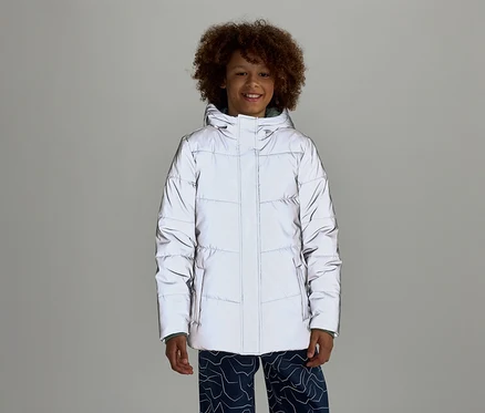 Un garçon souriant aux cheveux bouclés porte une doudoune réfléchissante à capuche et un pantalon bleu à motifs sur un fond gris.