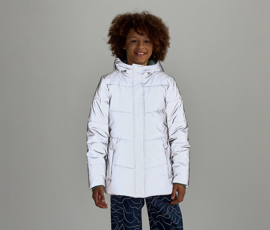Un garçon souriant aux cheveux bouclés porte une doudoune réfléchissante à capuche et un pantalon bleu à motifs sur un fond gris.