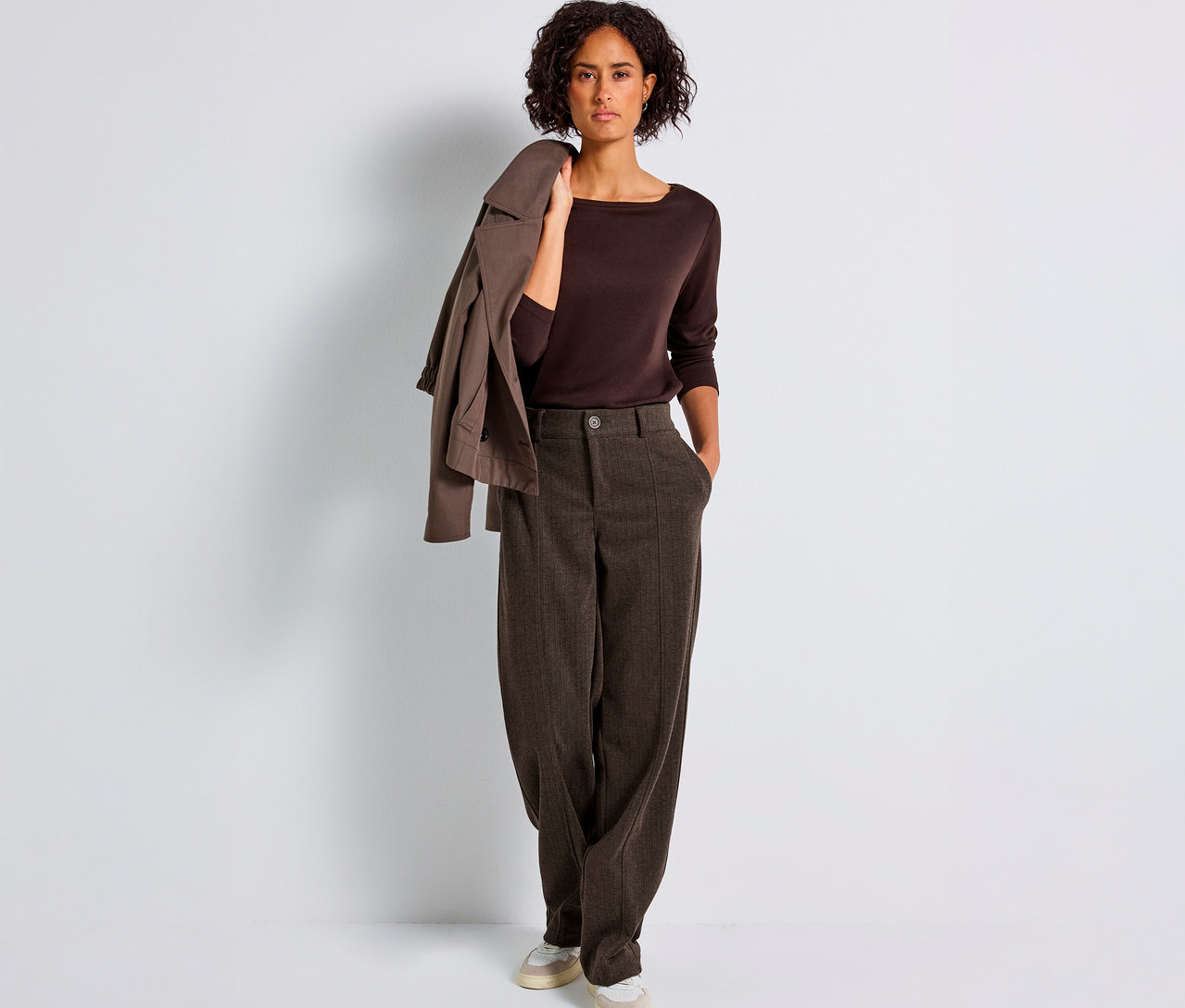Une femme debout porte un pantalon droit marron Street One, un haut marron et une veste sur l'épaule.