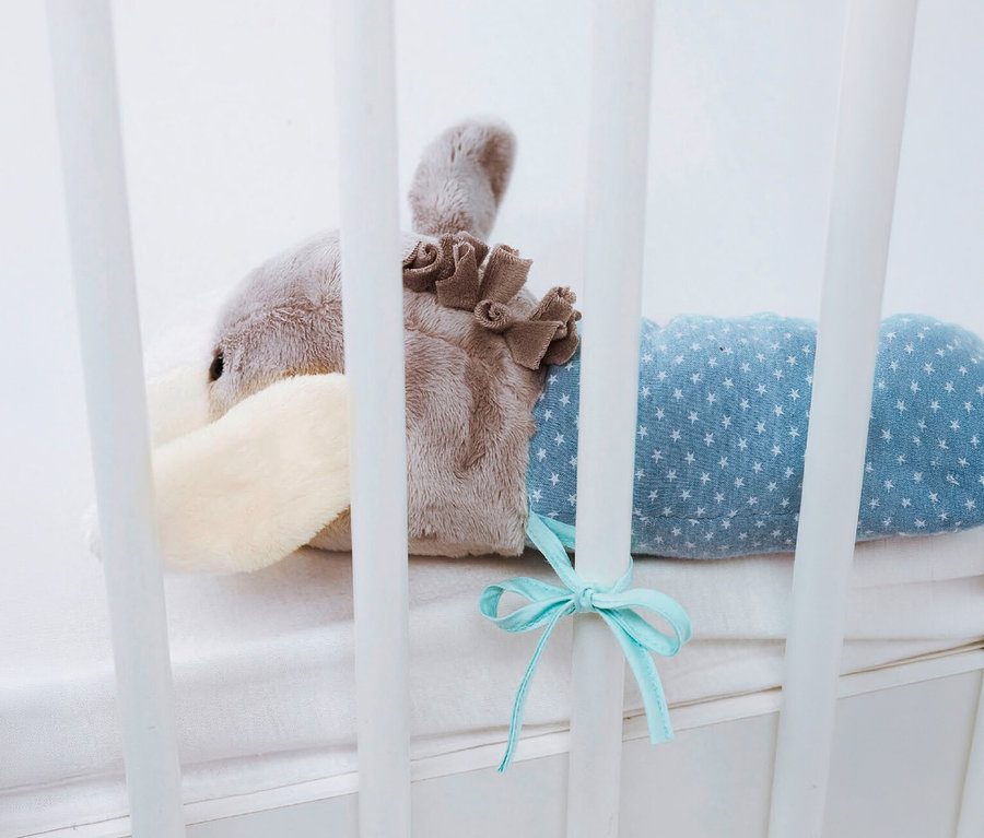 Un âne en peluche gris et crème avec un corps bleu à motifs d'étoiles, attaché aux barreaux blancs d'un lit de bébé.