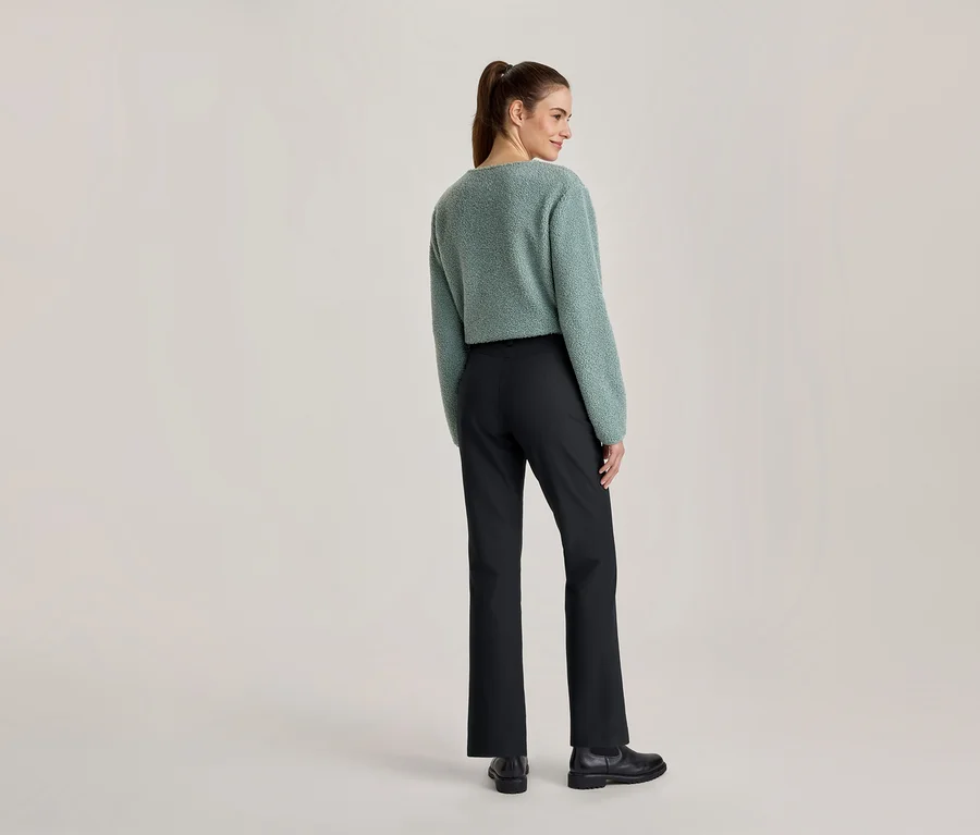 Femme vue de dos, légèrement tournée, portant un pull duveteux vert clair, un pantalon noir et des chaussures noires.