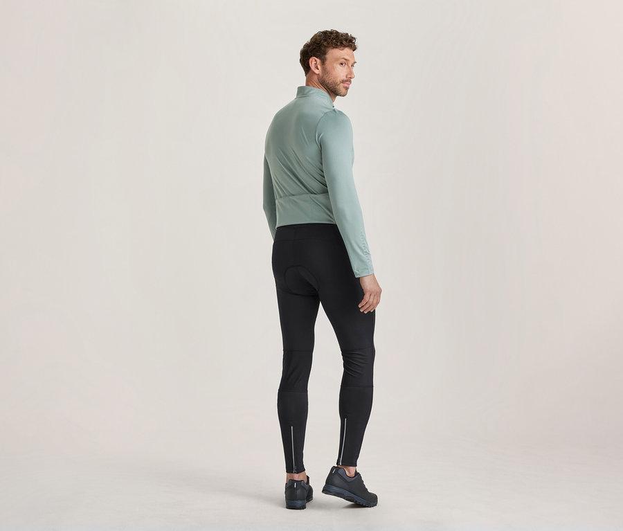 Homme debout de dos, regardant par-dessus son épaule droite, portant un haut à manches longues vert clair, un cuissard de cyclisme noir avec des fermetures éclair réfléchissantes aux chevilles et des chaussures foncées.