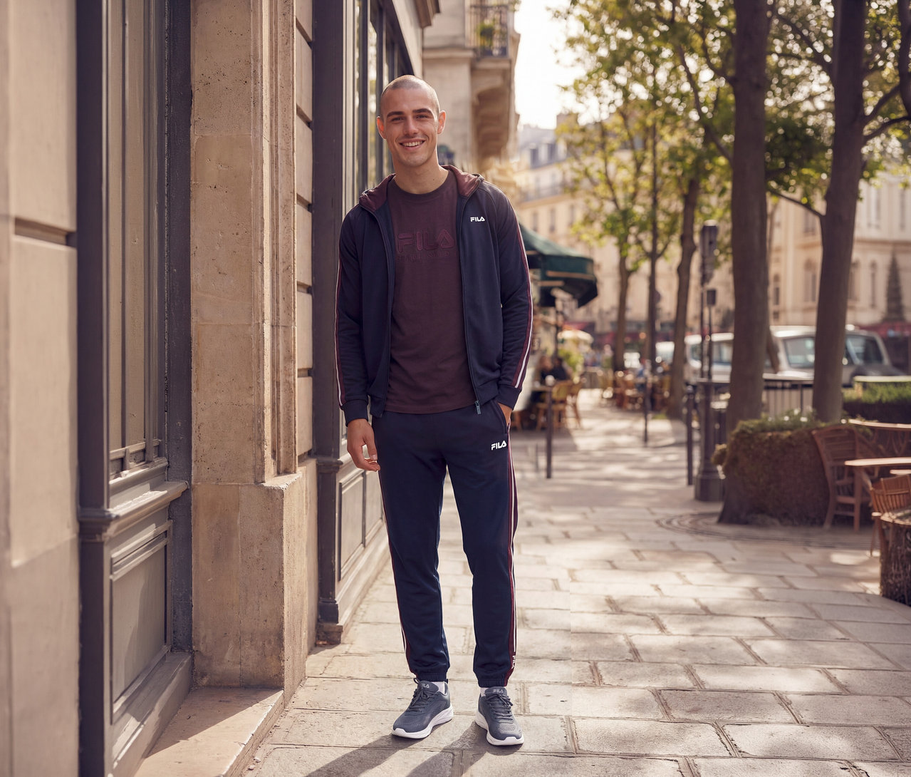 Homme souriant portant un survêtement bleu marine à rayures bordeaux, un t-shirt bordeaux et des baskets grises, debout sur un trottoir de ville.