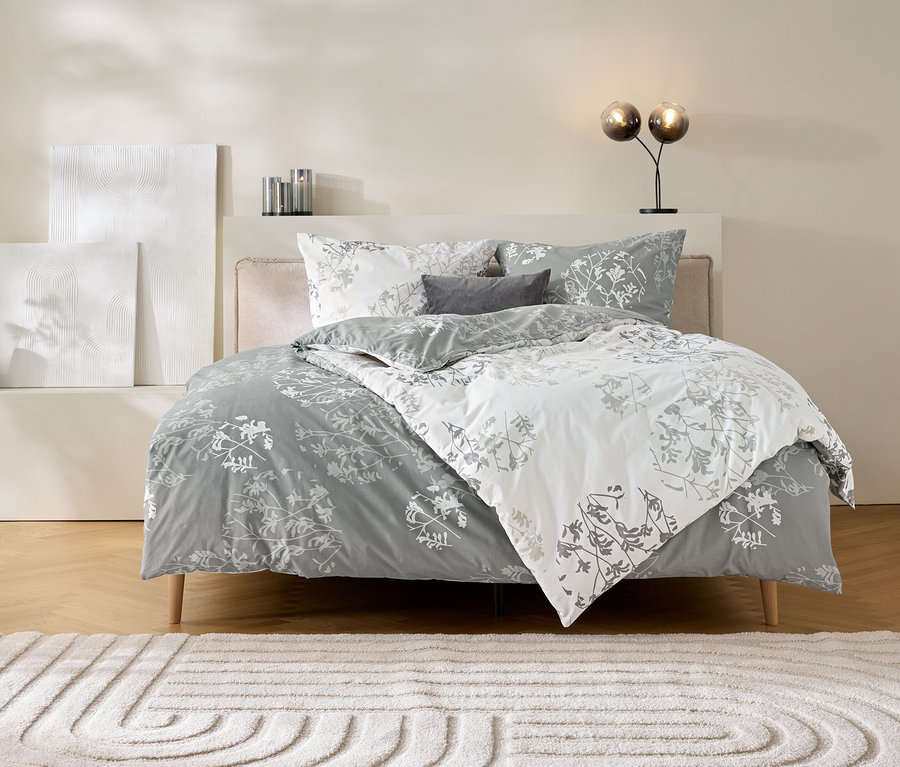 Un lit avec une parure de lit en renforcé grise à motif floral gris et blanc. La parure de lit est partiellement repliée, montrant les deux côtés. Deux oreillers et un petit coussin décoratif sont sur le lit. À gauche du lit, un vase noir avec des fleurs séchées et deux objets d'art blancs texturés. À droite, une lampe avec deux globes en verre. Devant le lit, un tapis clair avec un motif circulaire.