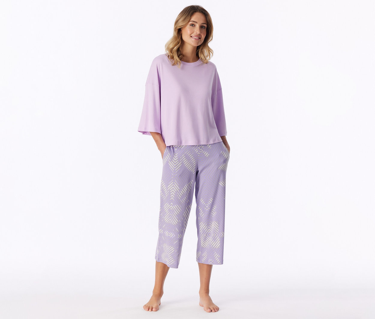 Femme souriante debout pieds nus, portant un haut à manches courtes violet clair et un pantalon capri violet clair à motif géométrique blanc, les mains dans les poches.