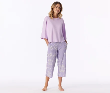 Femme souriante debout pieds nus, portant un haut à manches courtes violet clair et un pantalon capri violet clair à motif géométrique blanc, les mains dans les poches.