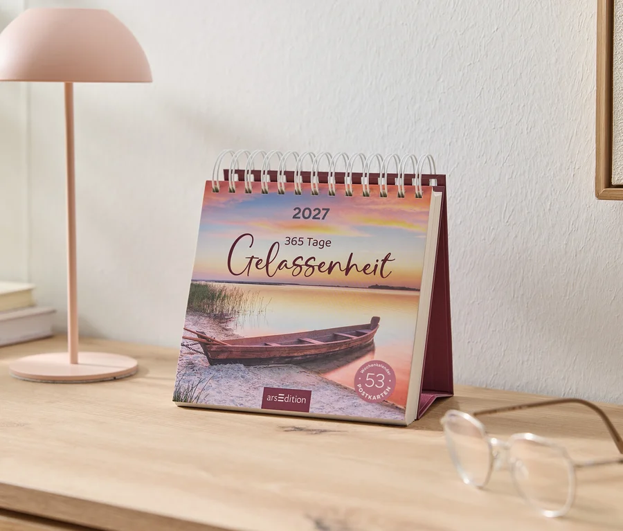 Un calendrier de bureau 2027 avec "365 Jours de Sérénité" et un bateau au bord d'un lac au coucher du soleil est posé sur une table en bois. À côté, une lampe rose, des livres et des lunettes.