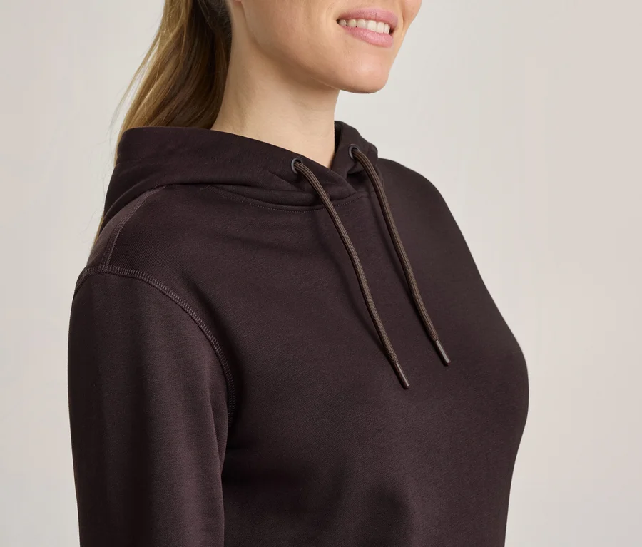 Femme en sweat à capuche marron foncé avec cordons, cheveux châtains en queue de cheval, souriante.