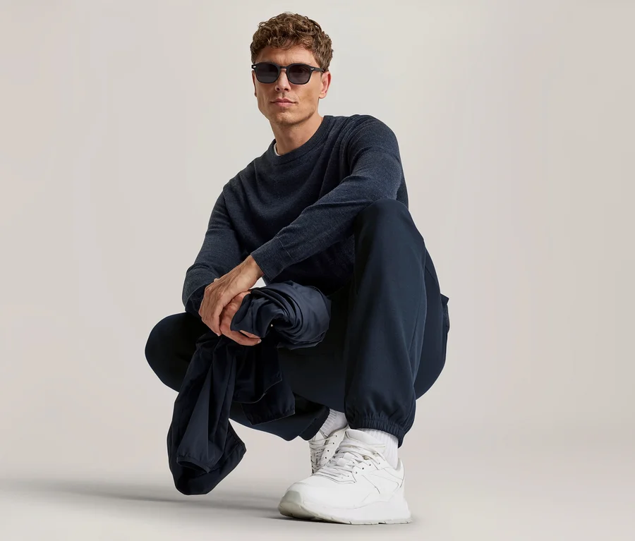 Homme aux cheveux bruns bouclés et lunettes de soleil accroupi, portant un haut à manches longues bleu foncé, un jogging bleu foncé et des baskets blanches, tenant un vêtement bleu foncé.