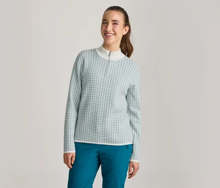 Une femme souriante regarde l'appareil photo. Elle porte un haut à manches longues à motif bleu clair et blanc, avec un col montant blanc et une courte fermeture éclair, et un pantalon bleu sarcelle foncé. Ses cheveux bruns sont attachés en queue de cheval.