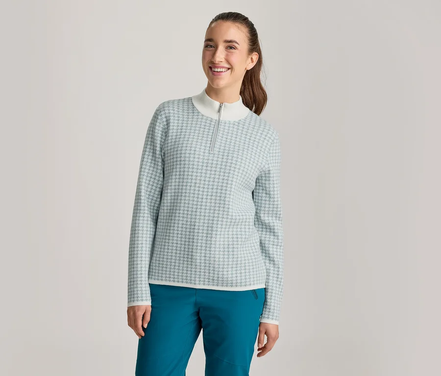 Une femme souriante regarde l'appareil photo. Elle porte un haut à manches longues à motif bleu clair et blanc, avec un col montant blanc et une courte fermeture éclair, et un pantalon bleu sarcelle foncé. Ses cheveux bruns sont attachés en queue de cheval.