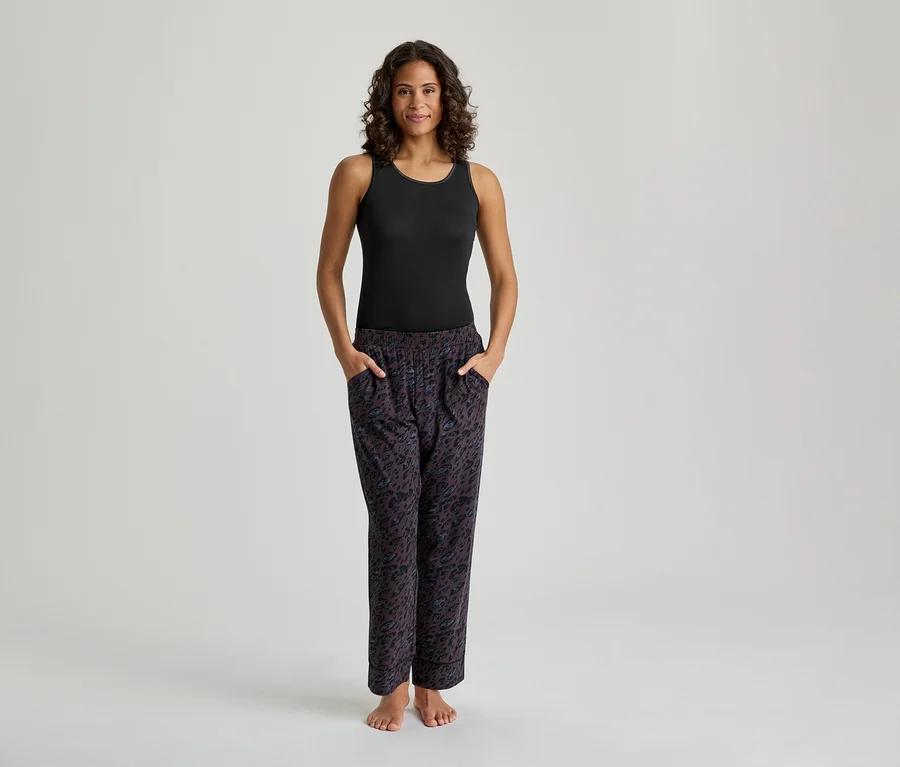 Une femme souriante se tient debout, vêtue d'un débardeur noir et d'un pantalon de pyjama à motifs, les mains dans les poches.