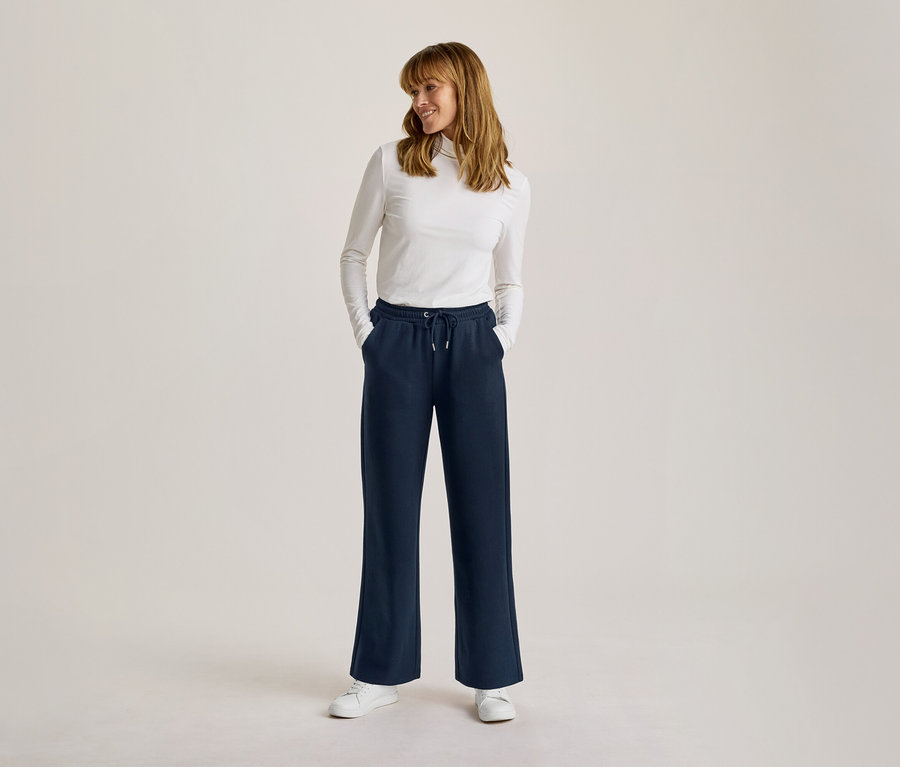 Une femme souriante se tient debout, les mains dans les poches d'un pantalon de jogging large bleu marine, avec un haut blanc à manches longues et des baskets blanches.