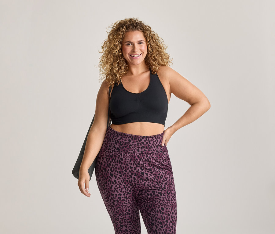 Femme souriante aux cheveux bouclés portant une brassière de sport noire et un legging violet à imprimé léopard, tenant un tapis de yoga sur son épaule.