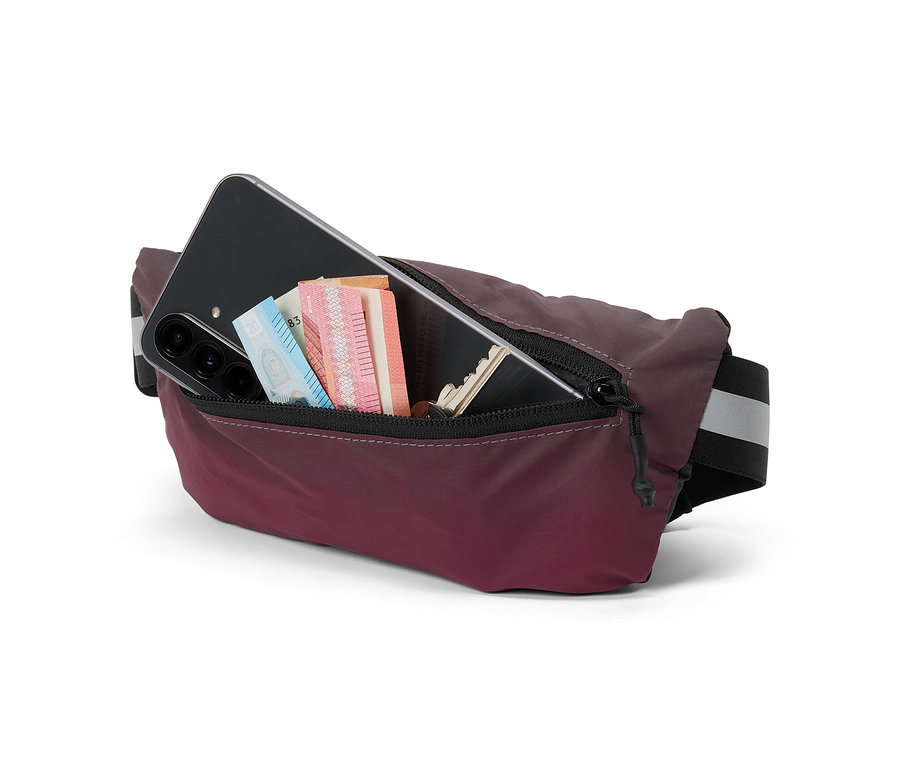Sac banane bordeaux avec fermeture éclair ouverte, contenant un smartphone noir, des billets de banque et une clé, sur fond blanc.