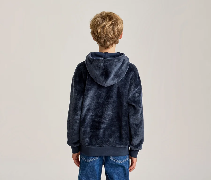 Une personne aux cheveux blonds courts et ondulés est vue de dos, portant un sweat à capuche bleu marine duveteux et un jean bleu, sur un fond clair et uni.