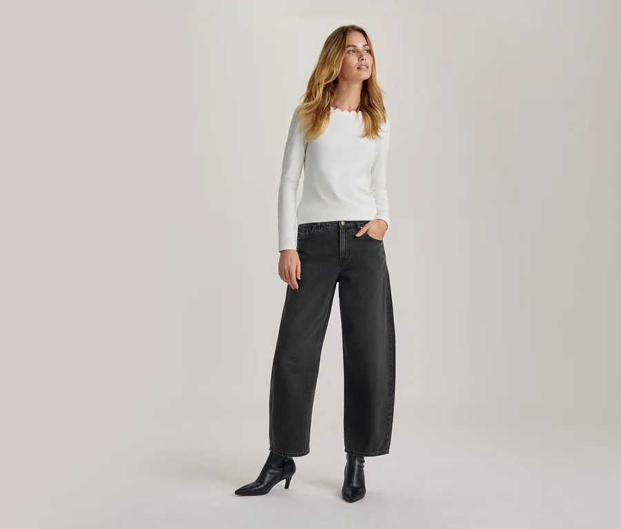Femme debout, portant un haut blanc à manches longues avec un col festonné, un jean large gris foncé et des bottines noires à talons. Sa main droite est dans sa poche, elle regarde vers la droite.