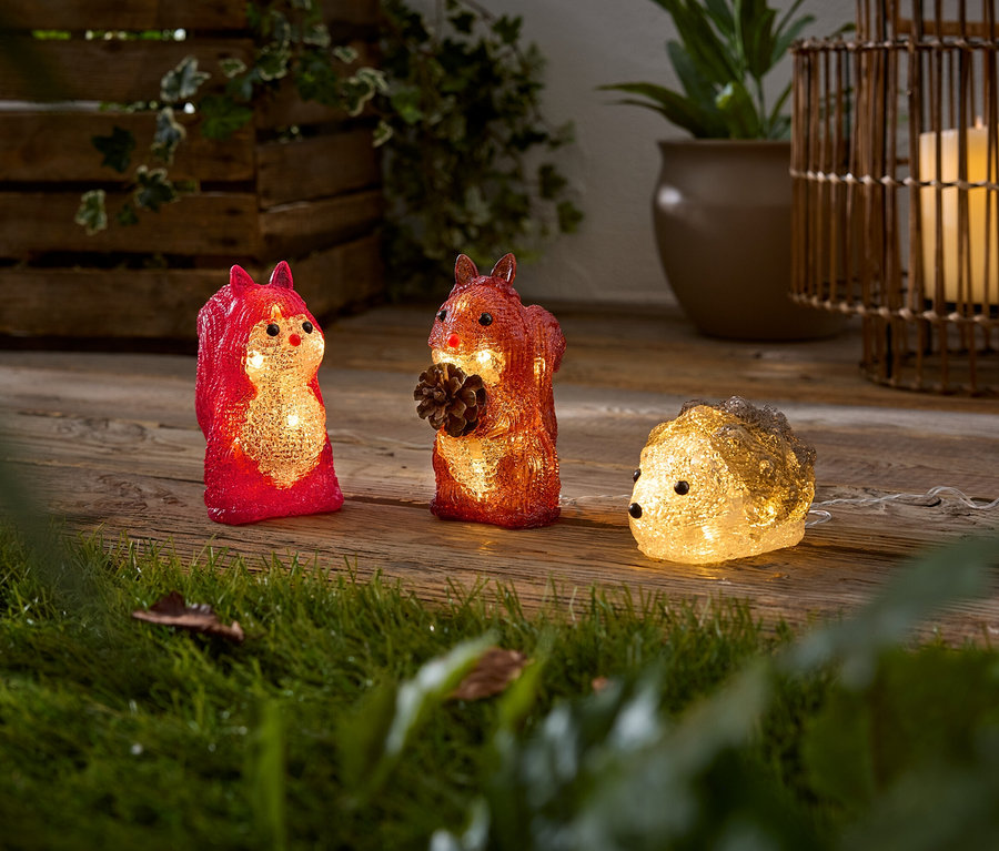 Trois figurines d'animaux lumineuses, un écureuil rouge, un écureuil orange tenant une pomme de pin et un hérisson jaune clair, sont posées sur une surface en bois. De l'herbe verte est au premier plan.