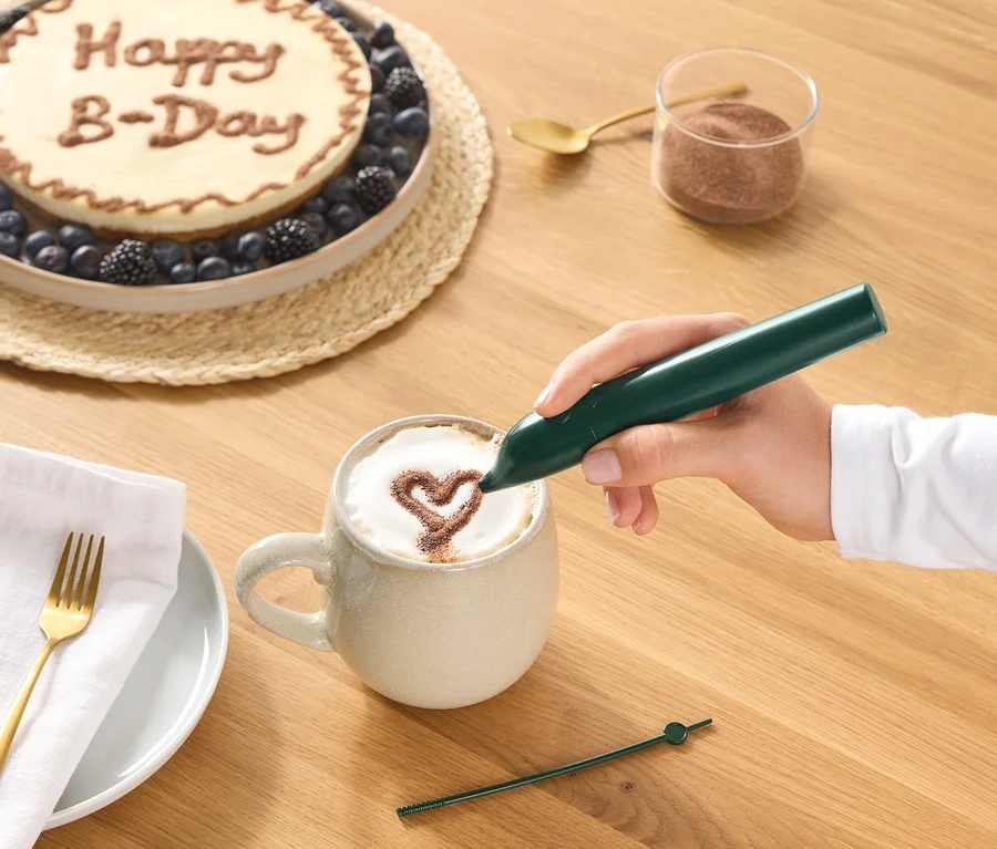 Une main dessine un cœur en cacao sur la mousse d'un café dans une tasse à l'aide d'un stylo décoratif vert. Sur la table en bois, on voit aussi un gâteau avec "Happy B-Day" et des baies, un verre de poudre de cacao, une cuillère dorée, et une assiette avec une serviette et une fourchette.