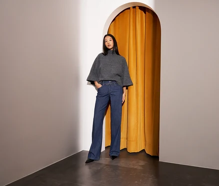 Femme debout dans un coin, portant un pull gris à col roulé et manches larges, et un pantalon large bleu à fines rayures, les mains dans les poches. Derrière elle se trouve une arche avec un rideau jaune.