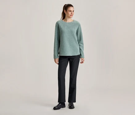 Femme debout portant un pull texturé vert clair, un pantalon noir et des chaussures noires, les cheveux attachés en queue de cheval.