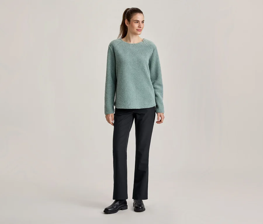Femme debout portant un pull texturé vert clair, un pantalon noir et des chaussures noires, les cheveux attachés en queue de cheval.