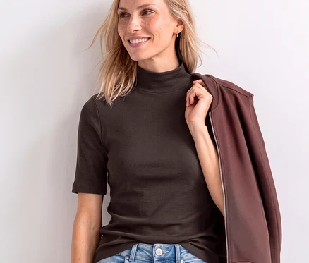 Femme souriante aux cheveux blonds portant un t-shirt Street One marron côtelé à col roulé et manches courtes, tenant une veste marron sur son épaule.