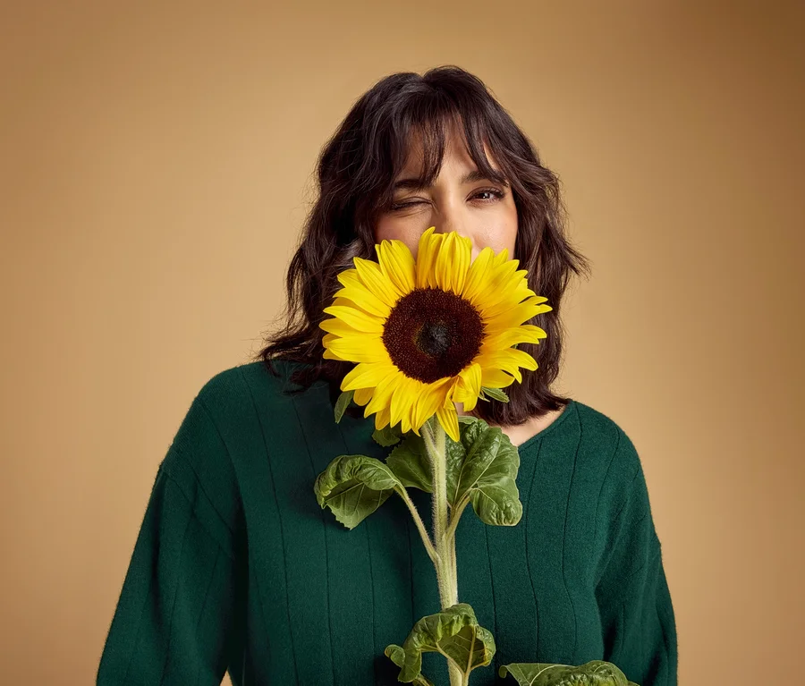 Une femme en pull vert foncé tient un grand tournesol jaune devant son visage, couvrant sa bouche et son nez. Elle cligne de l'œil visible.