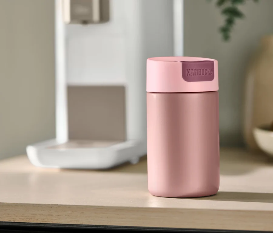 Mug de voyage rose Kambukka posé sur un comptoir en bois clair, avec une machine à café et des ustensiles de cuisine flous en arrière-plan.
