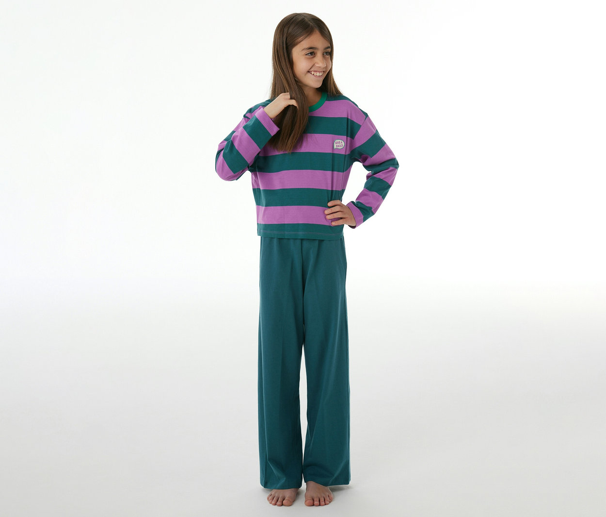 Jeune fille souriante, regardant à droite, vêtue d'un pyjama à manches longues rayé violet et vert et d'un pantalon large sarcelle, pieds nus.