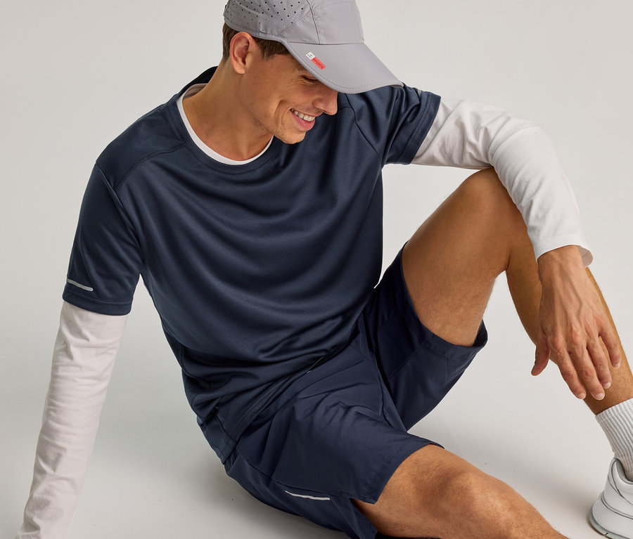 Un homme souriant est assis par terre, portant une casquette grise, un short de sport bleu marine et un t-shirt bleu marine sur un t-shirt blanc à manches longues.