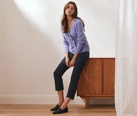 Une femme est assise sur une armoire en bois et porte un pantalon stretch 7/8 bleu foncé, un pull violet et des chaussures noires.
