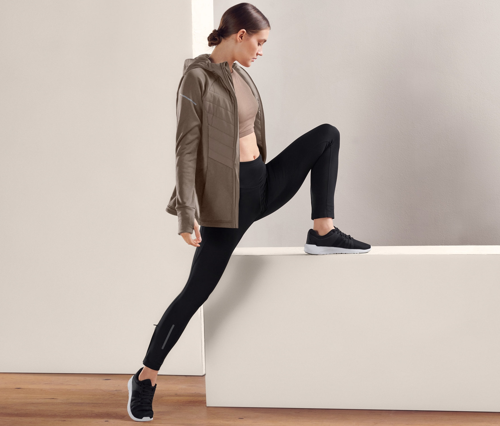 Une femme s'étire en portant un legging de sport haute performance noir, un soutien-gorge de sport et une veste marron.