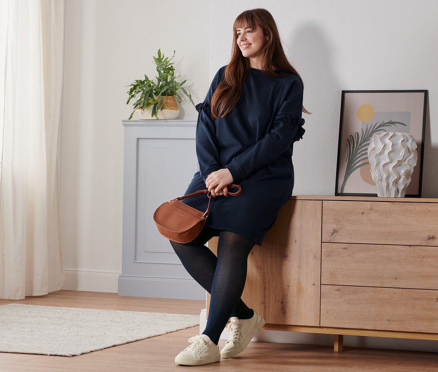 Femme assise sur une commode portant une robe molletonnée, des baskets en cuir et un sac à bandoulière.