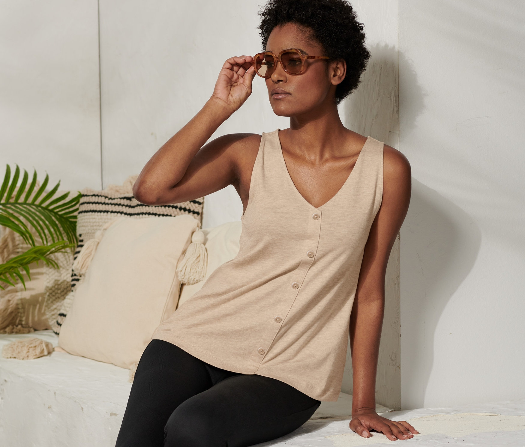 Femme assise ajustant ses lunettes de soleil. Elle porte un haut beige avec patte de boutonnage et un legging noir.