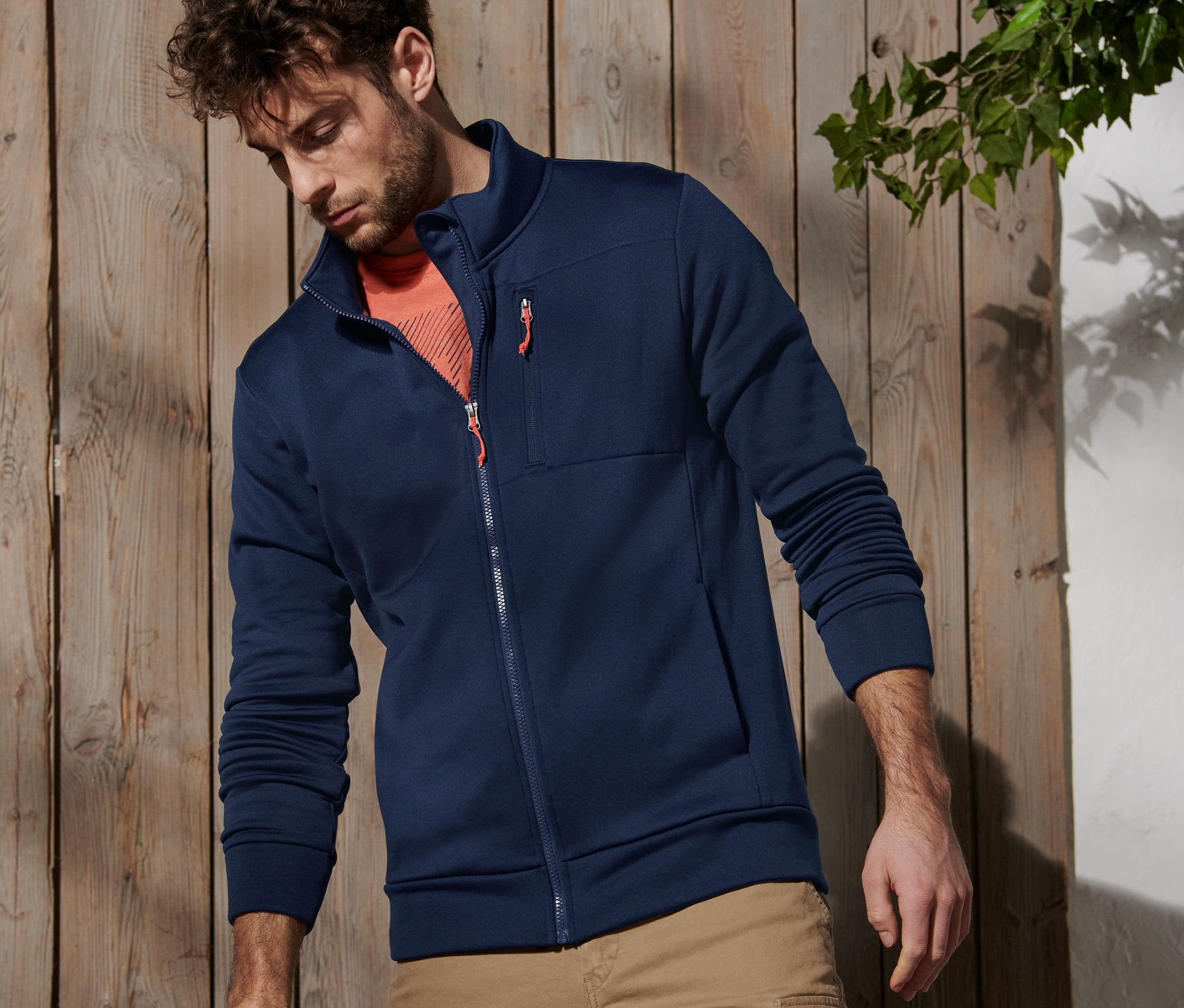 Un homme debout près d'un poteau en bois porte une veste fonctionnelle et un sac à dos est posé à côté de lui.