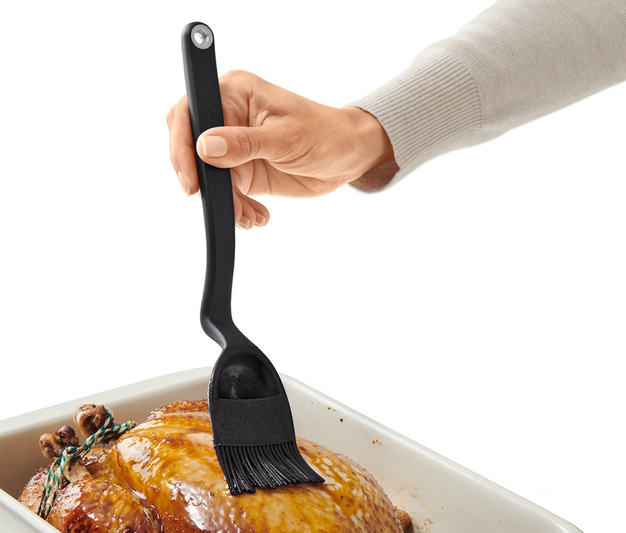 Une main badigeonne un poulet rôti dans un plat avec un pinceau à cuisson et à rôtissage.
