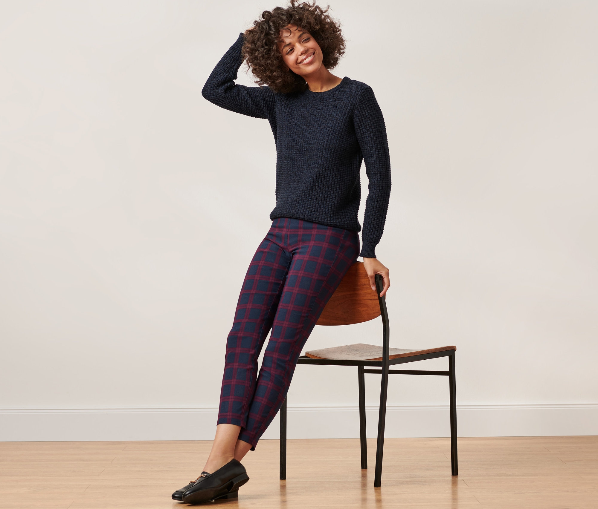 Une femme est assise sur une chaise et porte un pull bleu et un pantalon stretch à carreaux rouges et bleus.