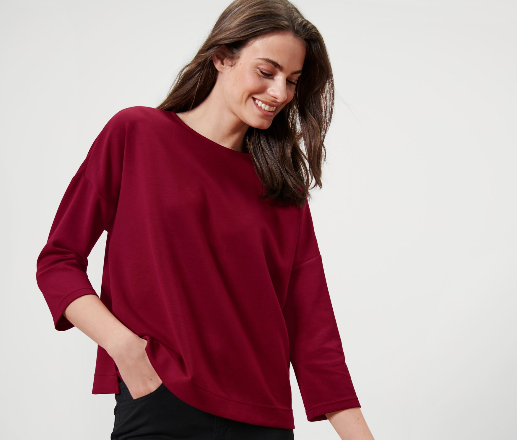Une femme debout, une main dans la poche de son pantalon, touchant une chaise avec l'autre main. Elle porte un sweat-shirt manches 3/4, rouge bordeaux.