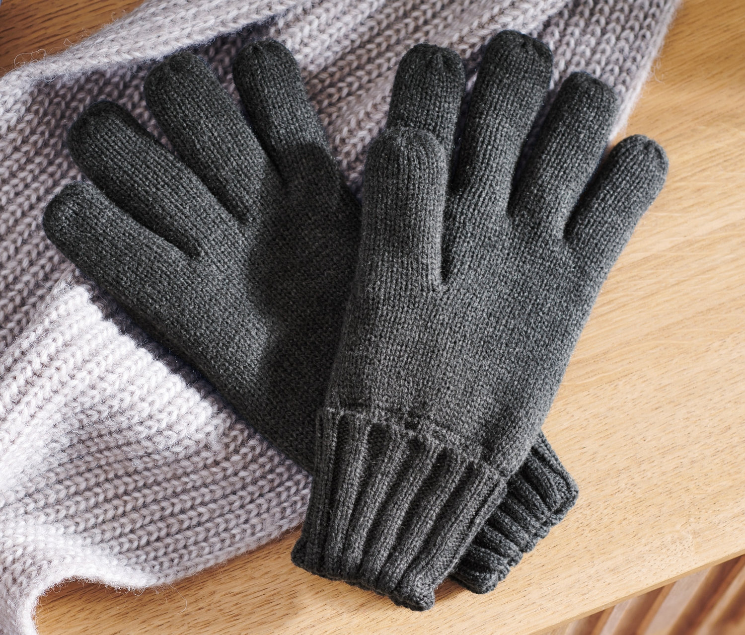 Des gants en maille gris reposent sur une écharpe en maille grise sur une surface en bois.