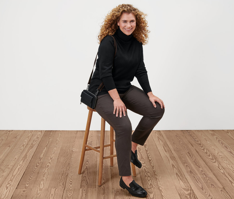 Une femme est assise sur un tabouret et porte un pull à col roulé en maille fine noir, un pantalon en bengaline marron, des mocassins en cuir et un mini sac.