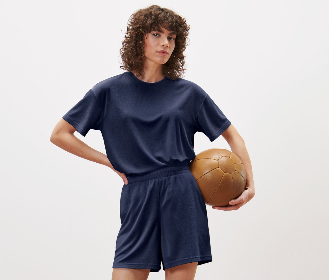 Femme posant en short de sport en tissu éponge bleu et t-shirt, tenant un ballon de football marron.